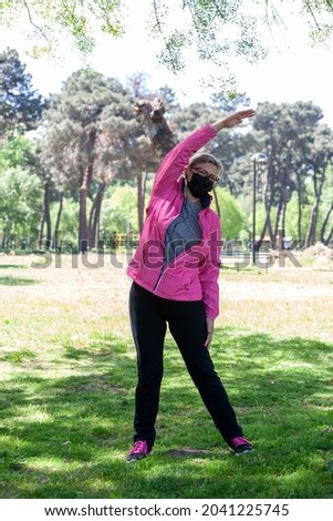 Mature woman trains in a park doing stretches while listening to music or some podcast with her wired headphones and wearing a surgical mask