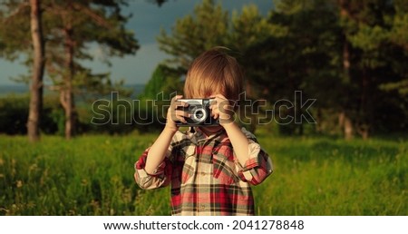 Waist up portrait view of the caucasian little boy smiling to the camera while taking a picture of green nature on a point-and-shoot digital camera. Happy childhood concept 