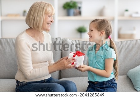 This Present Is For You. Portrait of loving girl sitting on couch greeting smiling senior lady with holiday or birthday, holding wrapped gift box, happy family celebrating special day together at home