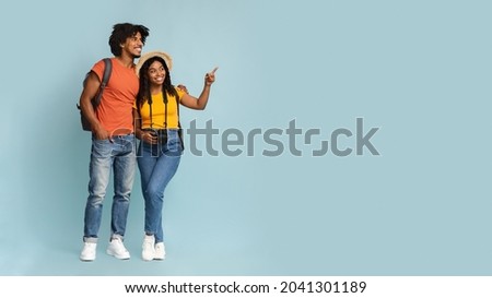 Happy millennial black couple tourists with backpacks and camera embracing and looking at copy space on blue studio background, choosing destination to travel, panorama. Travelling concept Royalty-Free Stock Photo #2041301189