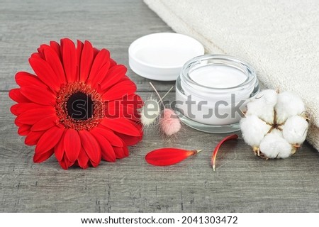Face cream jar on wooden background with red gerbera, soft face towel and cotton flower.