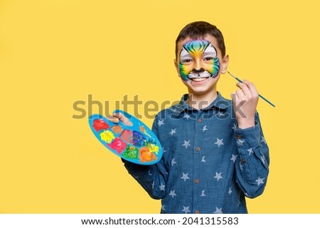 Portrait of ittle cute boy with faceart on birthday party, cute colorful tiger holding palette with gouache isolated on yellow background.