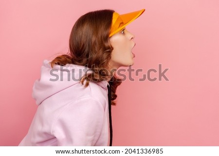Profile of surprised curly haired teenage girl in hoodie and yellow sun visor cap looking with shocked expression to side. Indoor studio shot isolated on pink background