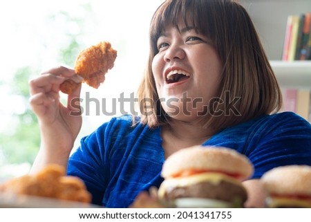 Hungry overweight woman holding Fried Chicken, hamburger on a wooden plate and Pizza on table, During work from home, gain weight problem. Concept of binge eating disorder (BED).