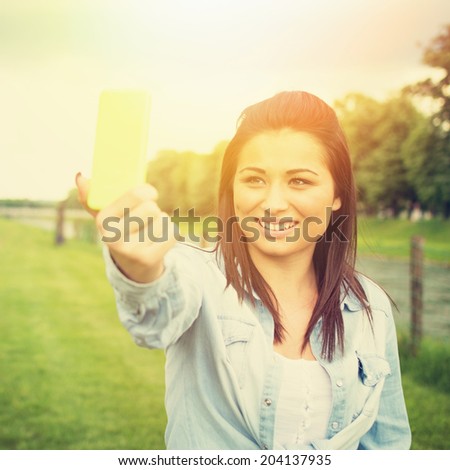 Happy young mixed race woman talking a selfie with smart phone outdoors in park on sunny summer day. Fashionable cute teenage girl photographing herself outdoors smiling wearing denim shirt.