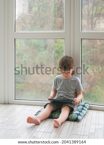 Curious boy watch cartoons on digital tablet. Kid sits on floor and uses electronic device. Indoor leisure for children while it's raining outside.