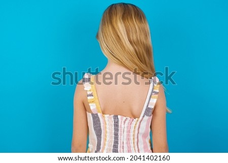 beautiful caucasian little girl wearing striped dress over blue background standing backwards looking away with arms on body.
