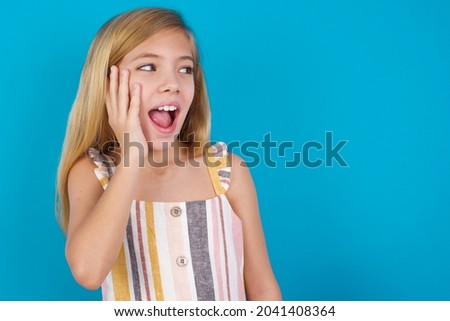 beautiful caucasian little girl wearing striped dress over blue background excited looking to the side hand on face. Advertisement and amazement concept.