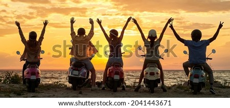 Back view of cheerful young friends, motorcycle riders raised their hands while standing with their scooters on the beach and watching sunset together. Friendship, nature concept. Rear view Royalty-Free Stock Photo #2041443671