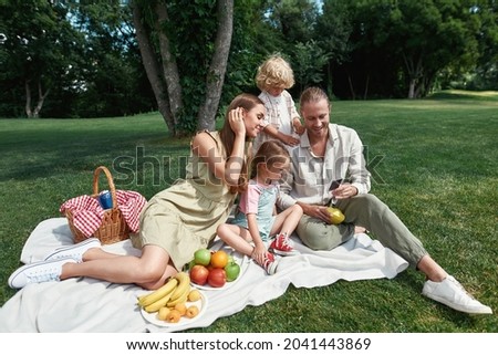 Lovely family spending time outdoors. Parents with little kids looking at photos they took while having picnic in nature on a summer day. Leisure, summer, technology concept