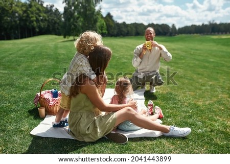 Happy family spending time outdoors. Mom with little kids posing while dad taking a picture using film camera in nature on a summer day. Leisure, summer, technology concept