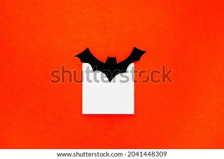 Crafts for the celebration of the concept of Halloween. Bat, cut out of paper holding a white square. Orange background. Copy space, place for text. Minimal Halloween background.
