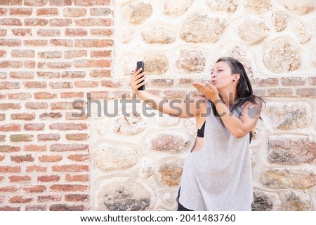 Young woman taking selfie photo by smart phone sending sweet air kiss, on wall background in city street, lifestyle concept with copy space for text.