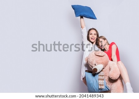 Pajama party. Meeting of friends. Two girls, one holding a pillow in her hand, lifting it to the top, the other laughing and holding a teddy bear. Girlfriends play with a plush toy.