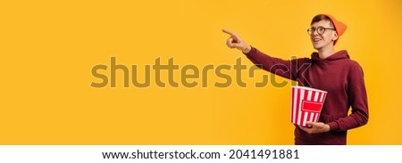 Photo of attractive crazy man holding big bucket of popcorn, eating popcorn, laughing out loud, on isolated yellow background