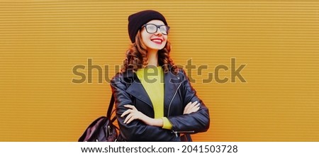 Portrait of stylish smiling young woman posing wearing backpack, black rock style leather jacket, hat, eyeglasses on orange background