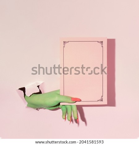 Creative layout with green painted hand holding pink paper copy space against pastel pink background. Halloween surreal idea. Minimal flat lay concept.