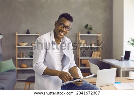 Portrait of confident happy African American man in his cozy and stylish home office. Relaxed confident handsome businessman in casual clothes holding notebook and looking at camera smiling.