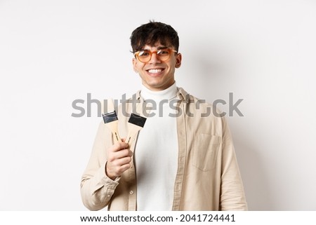 Hobbies and leisure concept. Stylish young man in glasses showing painting brushes and smiling, inviting to paint something, standing on white background