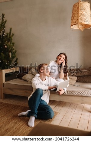A smiling young man and a woman in love are sitting on the sofa hugging working using wireless technologies looking at a laptop and a phone during the winter holidays in a cozy house
