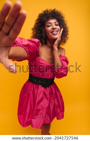 image of beautiful exotic appearance of girl in crimson dress on yellow background. curly brown-haired woman stretches her hand to camera smiling cute with teeth.