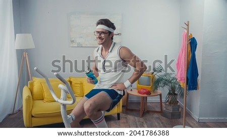 cheerful sportsman training on exercise bike and holding sports bottle in living room, retro sport concept