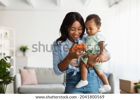 Happy young African American woman showing funny educational application on mobile phone to cute small son, lady holding baby on hands relaxing together, female using digital gadget, watching videos