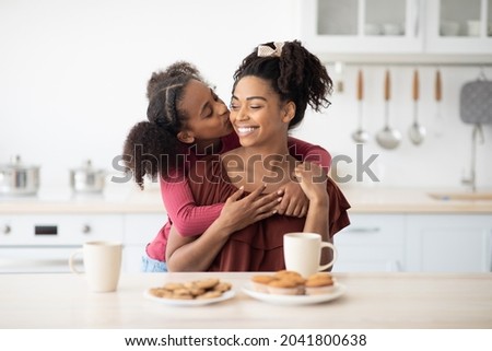 Cute black girl teenager hugging and kissing her happy beautiful mom after breakfast together, kitchen interior. Loving afro-american mother and daughter bonding while having snack, copy space