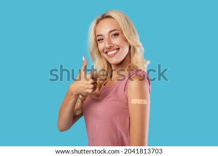 Successful Inoculation Against Coronavirus. Smiling Vaccinated Young Woman With Adhesive Bandage Gesturing Thumbs Up Showing Shoulder With Patch After Covid-19 Vaccine Injection, Blue Background Royalty-Free Stock Photo #2041813703