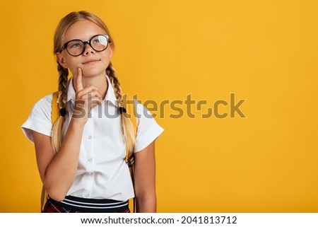 Pensive adolescence european cute schoolgirl with pigtails, backpack in glasses thinks and looks up at free space, isolated on yellow background. Creating plan and idea, female ponder about project