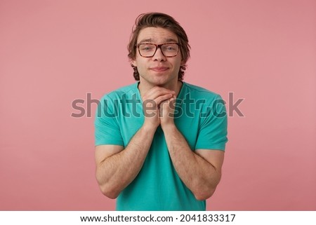 Indoor shot of young brunette male, keeps his arms together in praying position, keeps his eyes closed and think about exams result. Isolated over pink background
