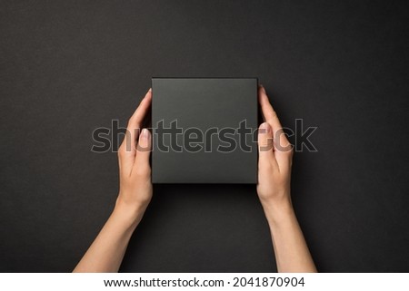 First person top view photo of hands holding black unwrapped giftbox on isolated black background with blank space