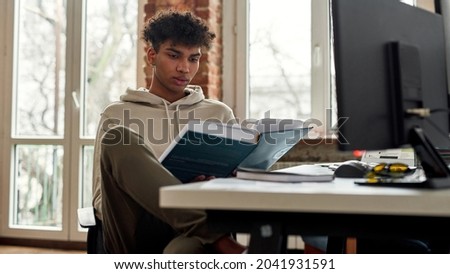 Millennial gen z African American male student read book at desk at home workplace. Focused young mixed race ethnicity man study work online on computer PC. Distant education, finance concept. Royalty-Free Stock Photo #2041931591