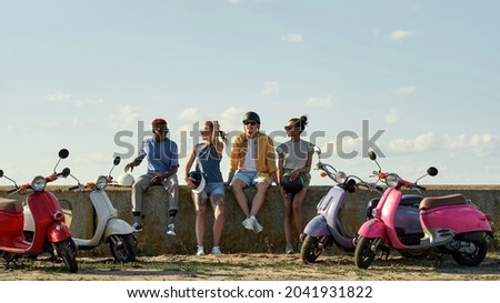 Cheerful young multiracial friends resting on concrete road fence beside retro scooters outdoors in warm sunny day, widescreen. Leisure, fun and entertainment concept Royalty-Free Stock Photo #2041931822