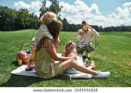 Back view of young mom with little kids posing while dad taking a picture of them using film camera in nature on a summer day. Leisure, summer, technology concept