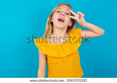 Caucasian blonde kid girl wearing yellow T-shirt against blue wall Doing peace symbol with fingers over face, smiling cheerful showing victory