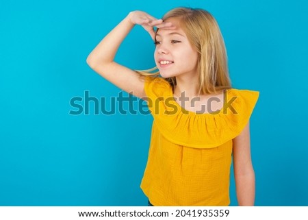 Caucasian blonde kid girl wearing yellow T-shirt against blue wall very happy and smiling looking far away with hand over head. Searching concept.