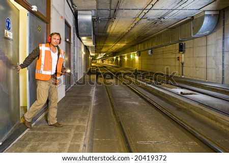 A sturdy looking engineer, closing a door to a maintenance room behind him inside a public transportation tunnel, looking relaxed and with a flashlight in his hand