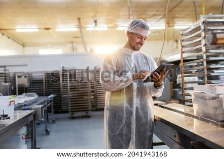 A happy food factory inspector in a sterile uniform is using a tablet to check on the quality and quantity of products. He is standing next to a line. He is very happy with the results.