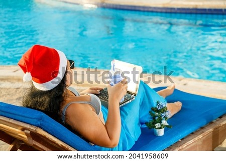 A woman in a Santa hat congratulates online friends on a sun lounger by the pool during the Christmas holidays next to a decorated Christmas tree. Horizontal photo.