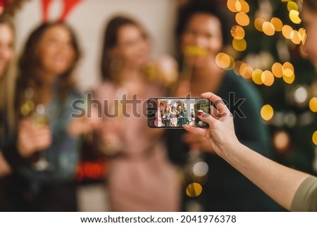 Group of cheerful multietnic female friends having fun and taking a memories photos with smartphone as they celebrate New Year at home together. Close-up of a smartphone.
