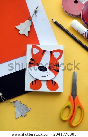 colored paper, tape, scissors, glue, pencil, box with a tiger applique, wooden Christmas trees on a yellow background. Children New year and Christmas Craft. Holiday gift wrapping vertical photo.
