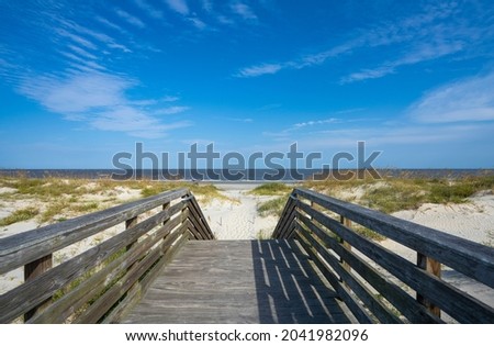 Pathway to the beach between grassy sand dunes. Wooden path over dunes at a beach leading to the ocean. Oceanview Beach Park, Jekyll Island, Georgia, USA. Royalty-Free Stock Photo #2041982096