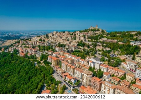 Aerial view of San Marino old town with old buildings and red roofs on the hill on a sunny day with clear sky. Picture from above with Italy behind in horizon