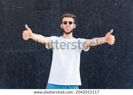 Gesture, emotion, expression and people concept - European bearded man in white T-shirt, glasses and jeans showing his tongue and thumbs up close-up in an open area on a black background 