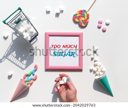 Text Too Much Sugar. Excess of sweets, chocolate, candy, sweet food in hand. Various tasty snacks in pastel colors, pink, mint green, on white background. Text in pink square frame. Sugar tax concept. Royalty-Free Stock Photo #2042029763