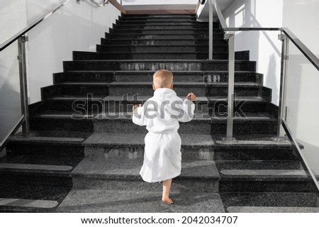 Back view of cute funny caucasian blond kid boy wearing white pool spa bathrobe walking upstairs on big marble staircase. Alone small child meet challenge and purpose. Rich luxury lifestyle concept Royalty-Free Stock Photo #2042034707