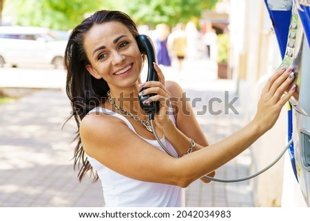 Positive brunette woman holding telephone receiver calls through stationery phone in street, tourist talking on payphone working with coins, happy with international communication on sunny summer day Royalty-Free Stock Photo #2042034983