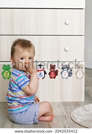 Thoughtful little baby boy with hand at mouth sitting in front of a chest of drawers with cartoon animals. Sweet 10 months old baby boy, getting to know animals, happy childhood and exploring concept.