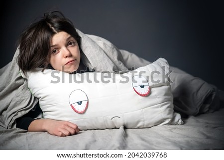 Girl hugs a pillow with tired red eyes and a sad expression on her face, hides under the covers. Woman is worried about insomnia, poor sleep, stress, in her hands a creative pillow with paper eyes.